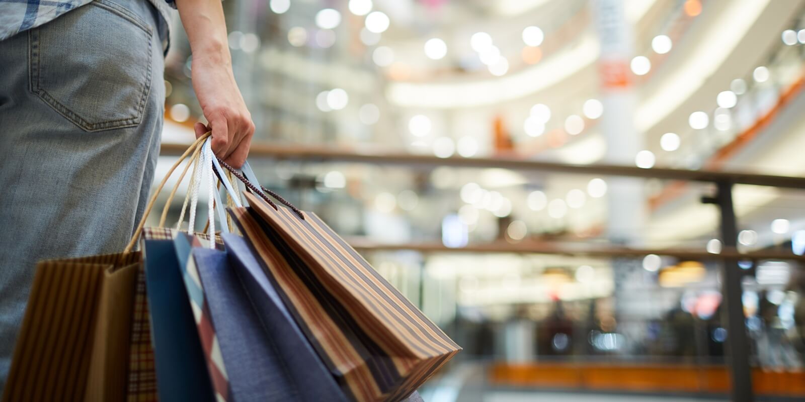 close-up of unrecognizable woman in casual clothing standing in spacious shopping mall with lights and holding many paper bags