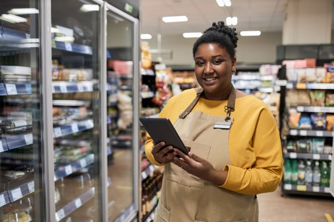 Waist up portrait of smiling black woman enjoying work in supermarket and looking at camera