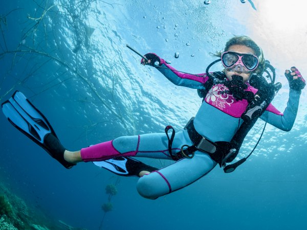 Happy little girl dives into the blue water of the ocean in scuba gear, mask and fins in the rays of sunlight, looking at the camera at the photographer. Lifestyle travel, beach adventure, summer vacation with baby, dive resort, kids diving, diving training