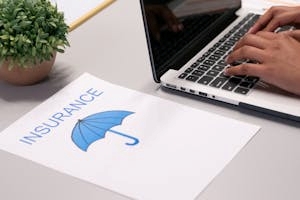 Person Working on Laptop Besides Paper with Text Insurance and Umbrella Image