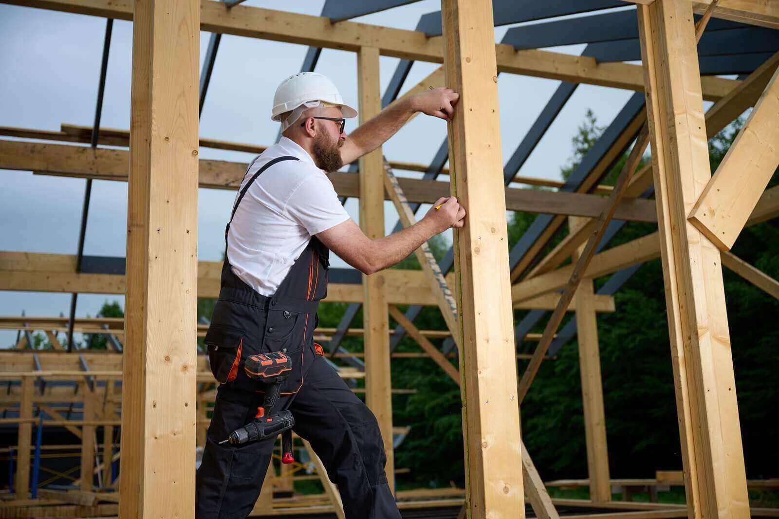 construction worker measuring wooden beam with tape measure at building frame