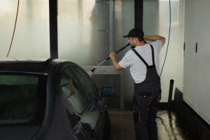 Seasonal Employee Carefully Washing and Cleaning a Dark Vehicle with a Powerful High-Pressure Water Sprayer