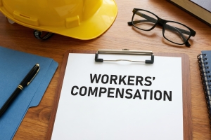 Document on Clipboard with Workers Compensation Text Near Contractor Hard Hat